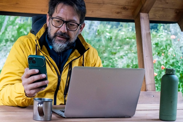 Photo one modern mature freelance using phone and laptop outdoor in forest cabin enjoying nature and freedom in business travel lifestyle adult man messaging outside wearing yellow jacket and glasses