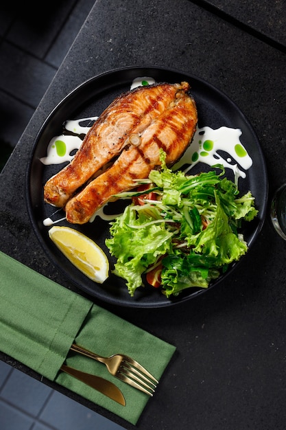 One piece of baked salmon grilled pepper lemon and salt on a black plate with lettuce leaves on a stone background