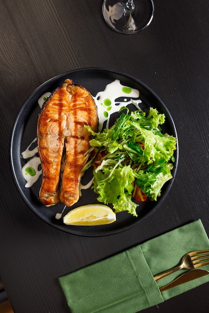 One piece of baked salmon grilled pepper lemon and salt on a black plate with lettuce leaves on a stone background