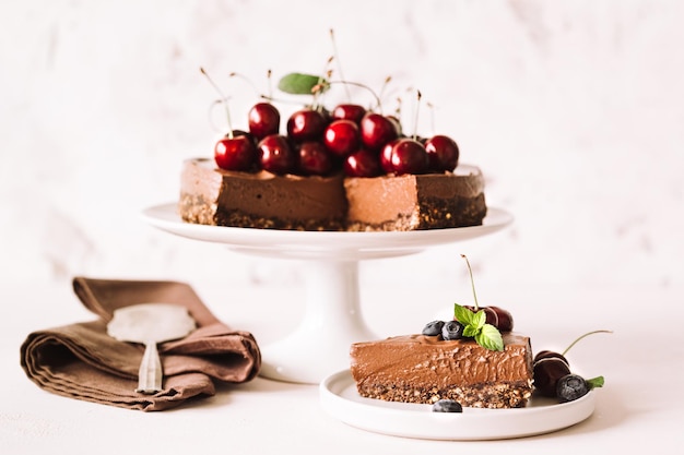 One piece of chocolate cheesecake decorated with fresh cherries, blueberries and a whole pie on a ceramic stand on a beige background. Horizontal.