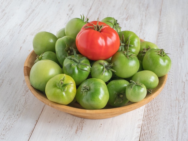 One red tomato among several green tomatoes on a wooden plate.