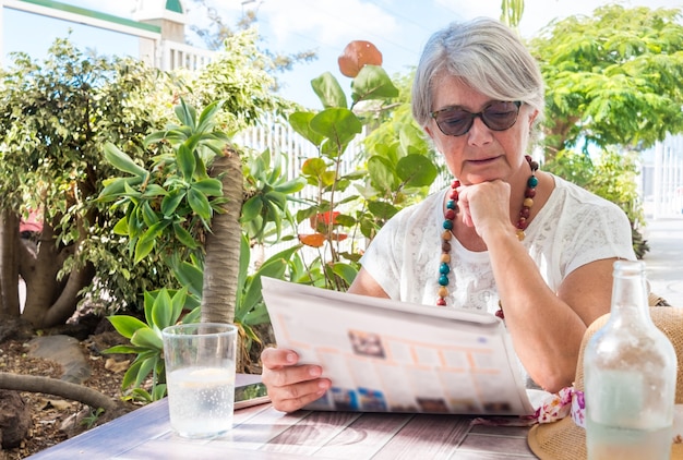 One retired pretty senior woman sitting and looking at the newspaper in the garden. Drinking a glass of water. Tropical plants and blue sky