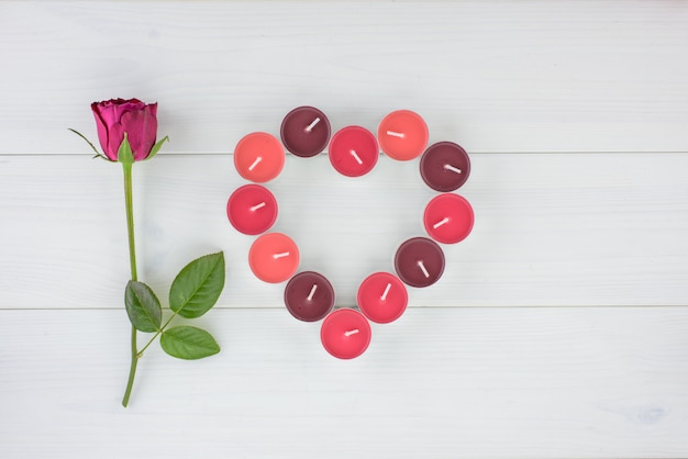 One strand of red rose and fragrant candles in heart shape on a white wooden table. 