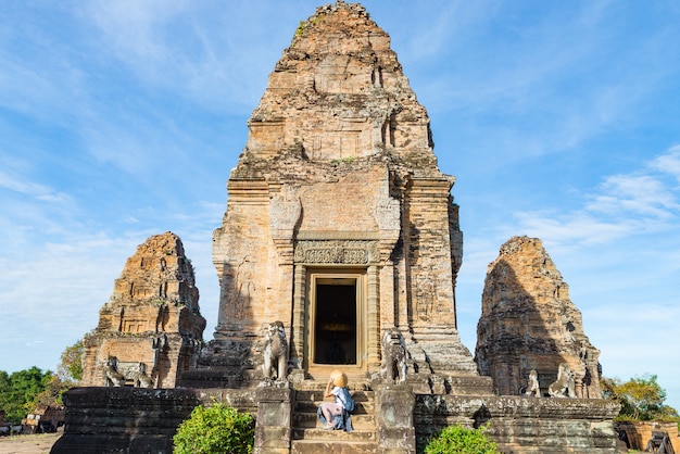 One tourist visiting Angkor ruins amid jungle, Angkor Wat temple complex, travel destination Cambodia. Woman with traditional hat, rear view.