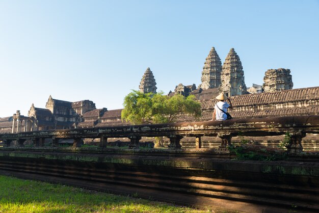 One tourist visiting Angkor ruins amid jungle, Angkor Wat temple complex, travel destination Cambodia. Woman with traditional hat, rear view.