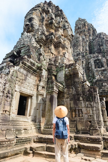 One tourist visiting Angkor ruins amid jungle, Angkor Wat temple complex, travel destination Cambodia. Woman with traditional hat, rear view.