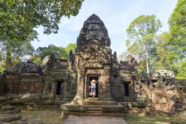 One tourist visiting Angkor ruins amid jungle, Angkor Wat temple complex, travel destination Cambodia. Woman with traditional hat, rear view.