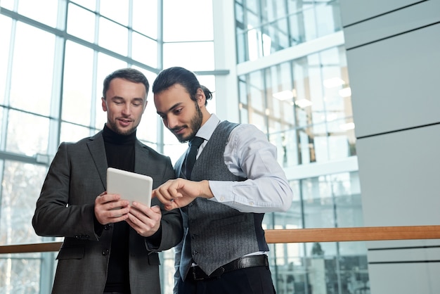 One of two young businessmen pointing at online document in tablet while explaining terms of electronic contract to colleague