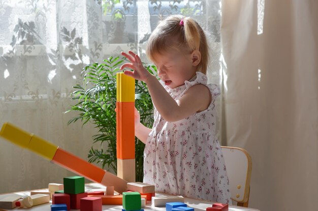 One twoyearold girl is sitting at a table and assembling a wooden construction kit A child breaks a tower of cubes