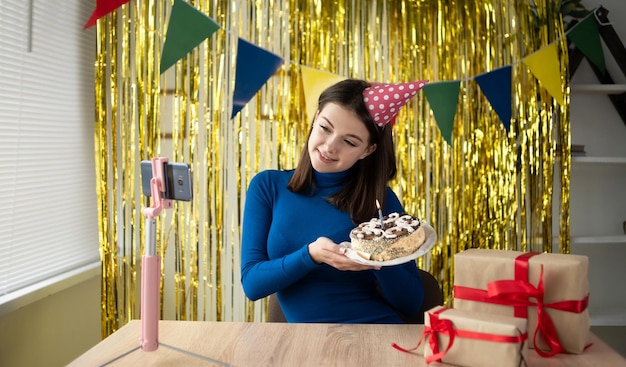 One woman celebrating her birthday alone at home with a video call and online conference via a smartphone using an application A lady in a festive cap on a yellow background holds a cake