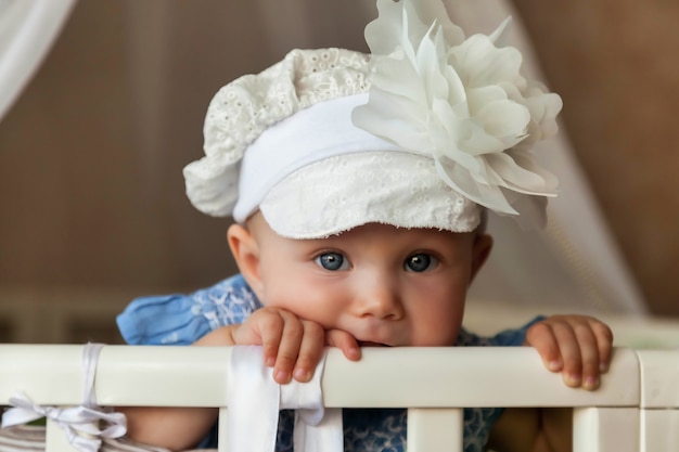 One-year-old blue-eyed child in stylish hat look at camera from crib. Small and cute baby with smile sitting in children's room and waiting mom. Concept of proper upbringing and childhood