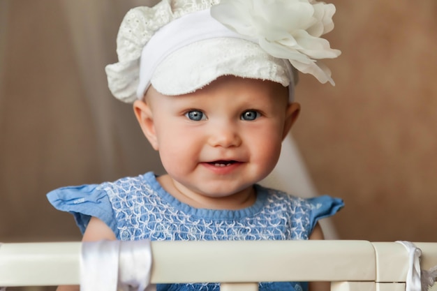 One-year-old blue-eyed child in stylish hat look at camera from crib. Small and cute baby with smile sitting in children's room and waiting mom. Concept of proper upbringing and childhood