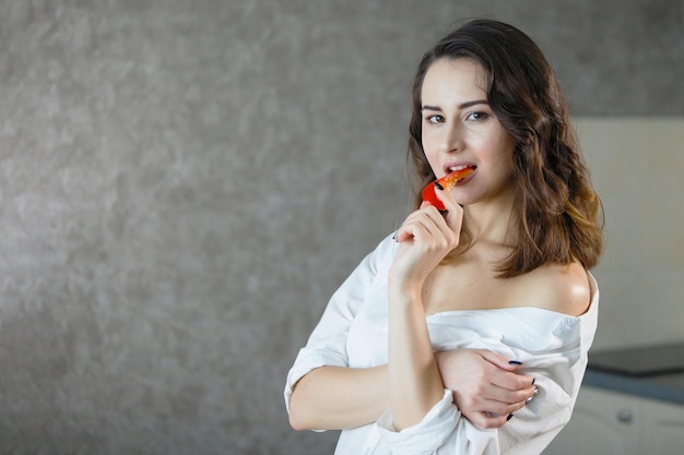 One young pretty woman in a white shirt in the kitchen is eating red pepper and smiling