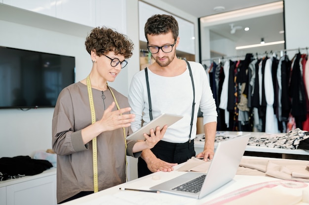 One of young tailor with tablet making presentation of ideas for new fashion collection while both standing by desk