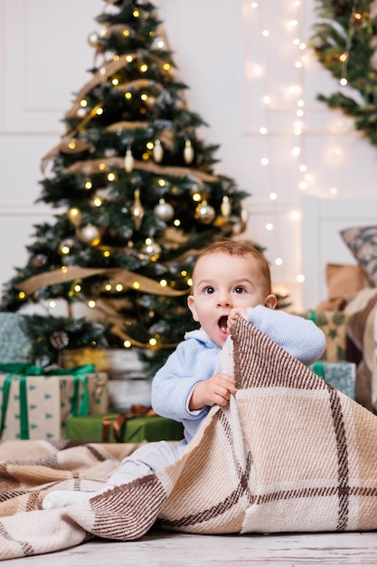 A oneyearold boy sits near a decorated Christmas tree with gifts Christmas tree in the house A happy child is waiting for the new year