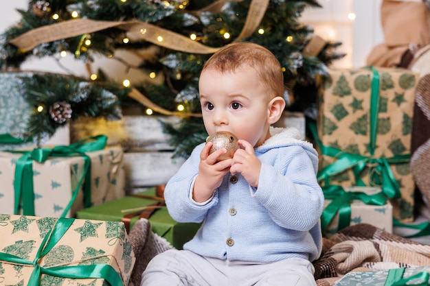 A oneyearold boy sits near a decorated Christmas tree with gifts Christmas tree in the house A happy child is waiting for the new year