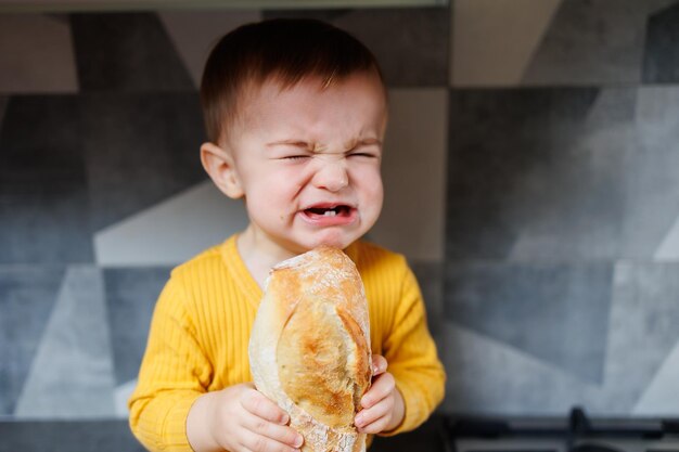 A oneyearold little boy in yellow clothes sits and eats freshly baked rye bread The child holds a fresh baguette in his hands