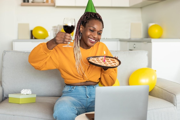 Online birthday Happy black woman celebrating bday showing pie with candle and glass of wine to laptop webcamera