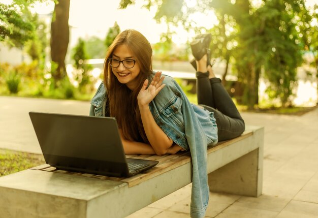 Online call. Distance learning. A young woman student in a denim jacket and glasses looks at a laptop screen while lying on a bench in a park