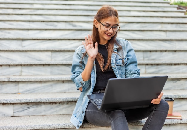 Online call. Young hipster woman in denim jacket communicates with friends on online conference with laptop while sitting on the stairs in the city