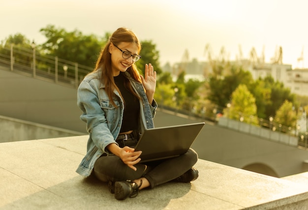 Online call. Young woman student in a denim jacket makes online broadcast while sitting on the stairs in the city outdoors. Distance learning