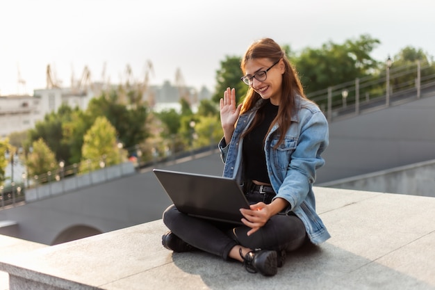 Online call. Young woman student in a denim jacket makes online broadcast while sitting on the stairs in the city outdoors. Distance learning