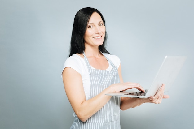 Online communication. Delighted woman keeping smile on face and holding laptop while standing in semi position over grey background