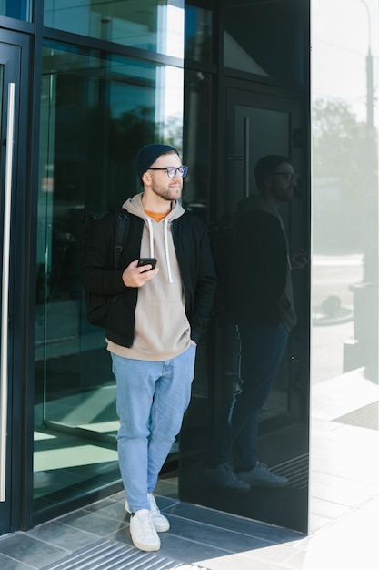 Online communication A young man in a hat and glasses with a smartphone in his hand on the street A young bearded guy on an urban background is chatting on a mobile phone