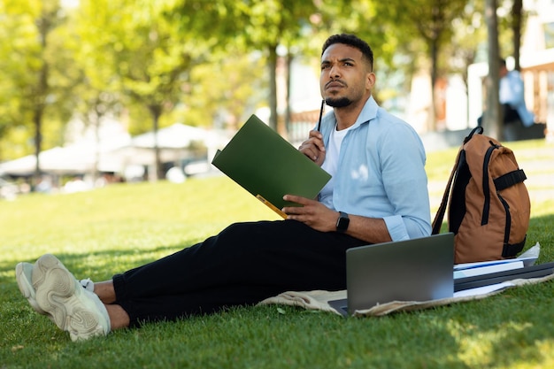 Online education concept Thoughtful african american male student studying outdoors using laptop and taking notes