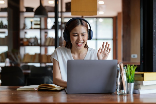 Online education A female student with headphones learning online at home she waving hand to other students or colleagues on computer monitor in video conference and smiling