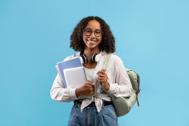 Online education smiling black female student with backpack and headphones holding notebooks and