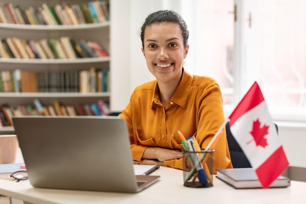 Online foreign languages tutoring Happy female teacher sitting in library with flag of Canada using laptop