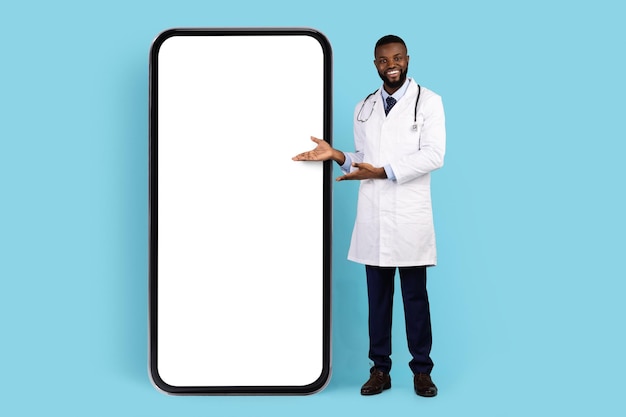 Online offer black smiling doctor in white robe pointing at blank smartphone