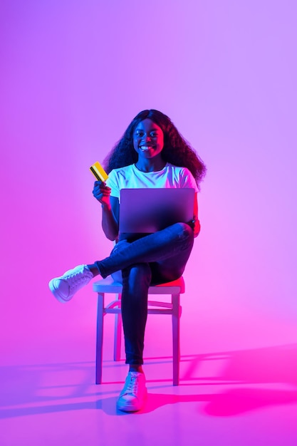 Online payments happy african american woman with credit card and laptop sitting on chair in neon