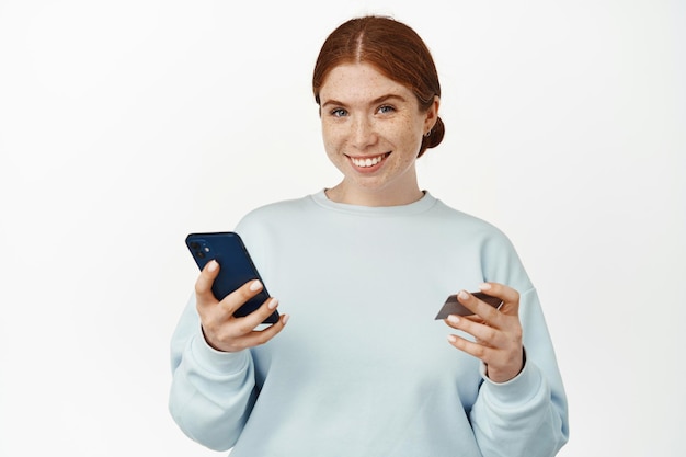 Online shopping. Beautiful young woman shops on website, app, paying with mobile phone and credit card, smiling confident and happy, standing against white background