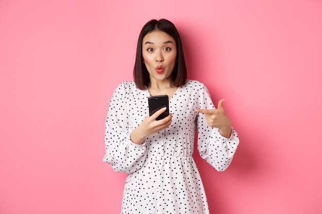 Online shopping and beauty concept. Amazed asian woman pointing finger at mobile phone, recommending smartphone app, standing over pink.