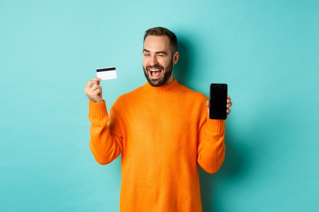 Online shopping.  caucasian man showing credit card and smartphone, winking at camera