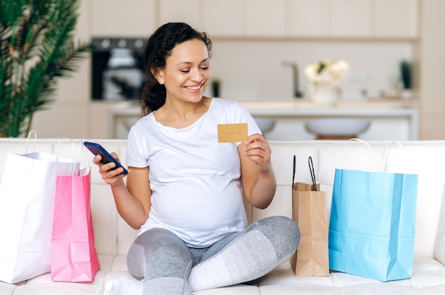 Online shopping concept Attractive successful happy pregnant mixed race woman sitting on the couch shopping online holding credit card and mobile phone making online payment smiling