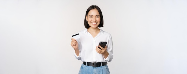 Online shopping concept Image of young asian modern woman holding credit card and smartphone buying with smartphone app paying contactless standing over white background