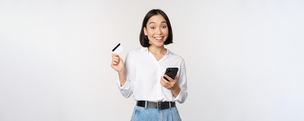 Online shopping concept Image of young asian modern woman holding credit card and smartphone buying with smartphone app paying contactless standing over white background