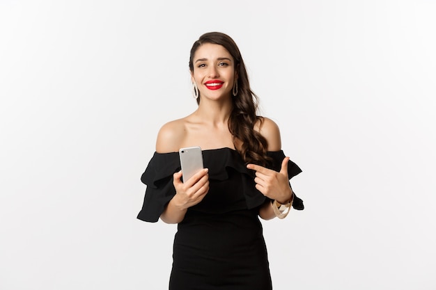 Online shopping concept. Stylish woman in black dress, wearing makeup, pointing finger at mobile phone with satisfied smile, standing over white background