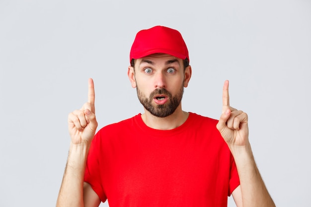 Photo online shopping, delivery during quarantine and takeaway concept. surprised and impressed delivery guy, courier in red t-shirt and cap, pointing fingers up, gasping amazed, read interesting info