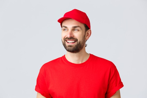 Online shopping, delivery during quarantine and takeaway concept. Cheerful bearded guy in red uniform cap and t-shirt, looking away with pleased smile, reading banner sign, grey background