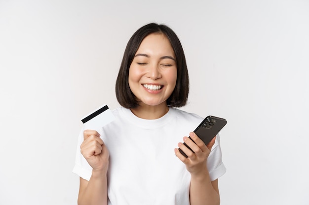 Online shopping Happy asian woman using credit card and smartphone app paying on website via mobile phone white background