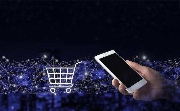 Online shopping, online store application in a smartphone. Hand hold white smartphone with digital hologram cart sign on city dark blurred background. The concept of Innovation in e-Commerce.