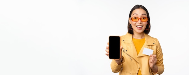 Online shopping and people concept Stylish asian woman showing mobile phone screen and credit card smartphone application standing over white background