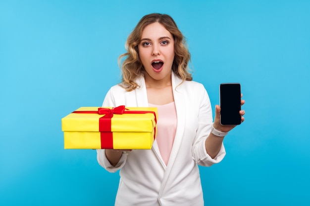 Online shopping Portrait of amazed beautiful woman with wavy hair in white jacket holding present box and phone looking with surprise at camera indoor studio shot isolated on blue background