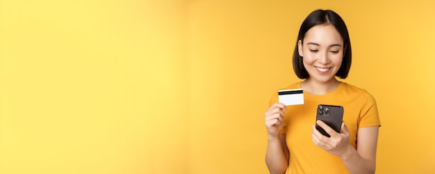 Online shopping Smiling asian girl using credit card and mobile phone app paying contactless order on smartphone application standing over yellow background