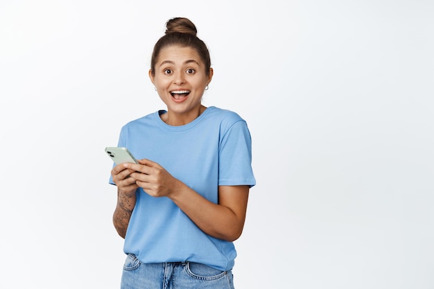 Online shopping and technology. Young woman smiling, holding mobile phone, wearing blue t-shirt, standing against white background.