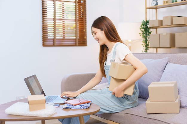 Online trader concept Female entrepreneur holds parcel boxes and checks online orders on laptop